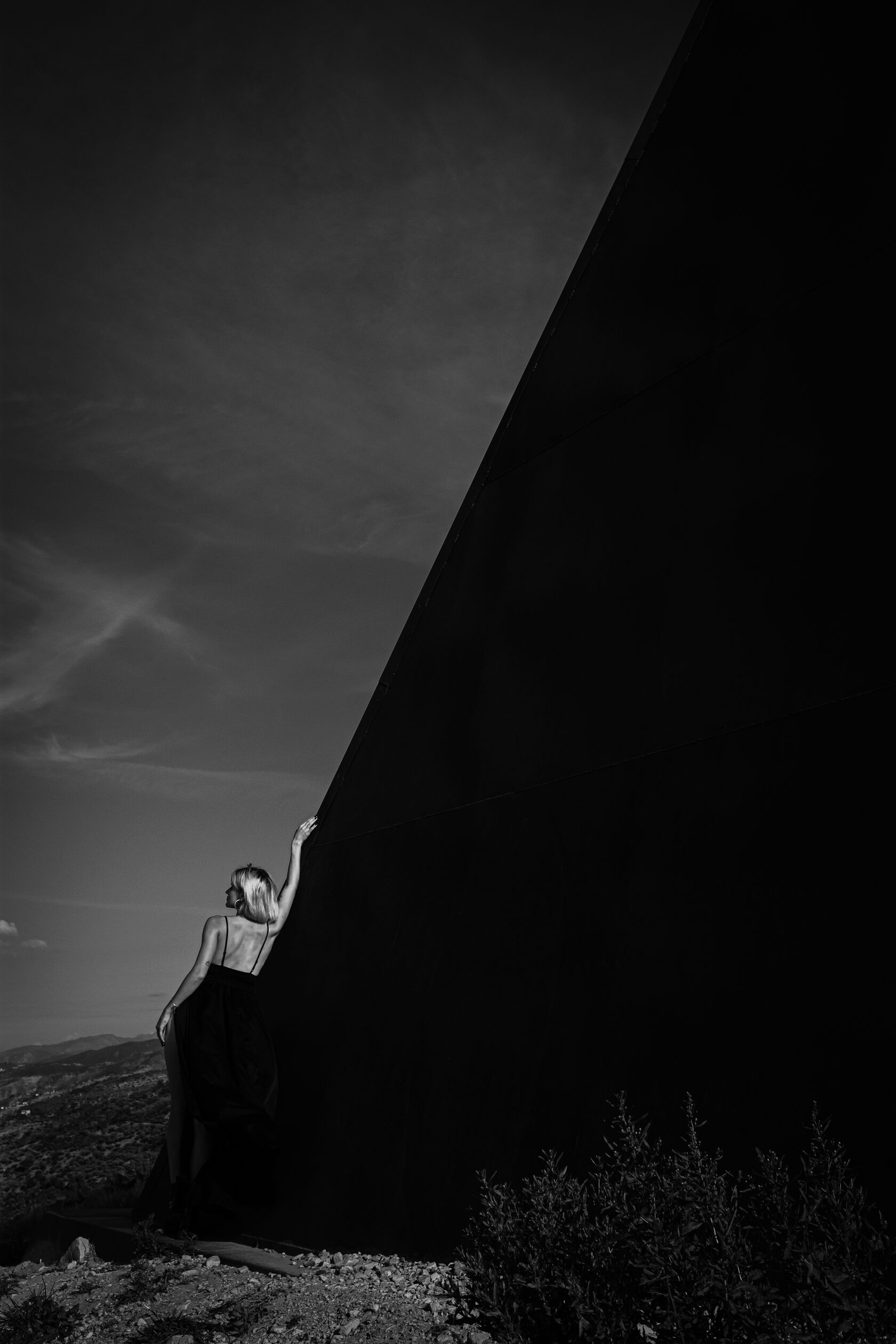 Silhouette of a person in a flowing dark dress touching a large dark angular structure against a pale sky, dramatic monochrome