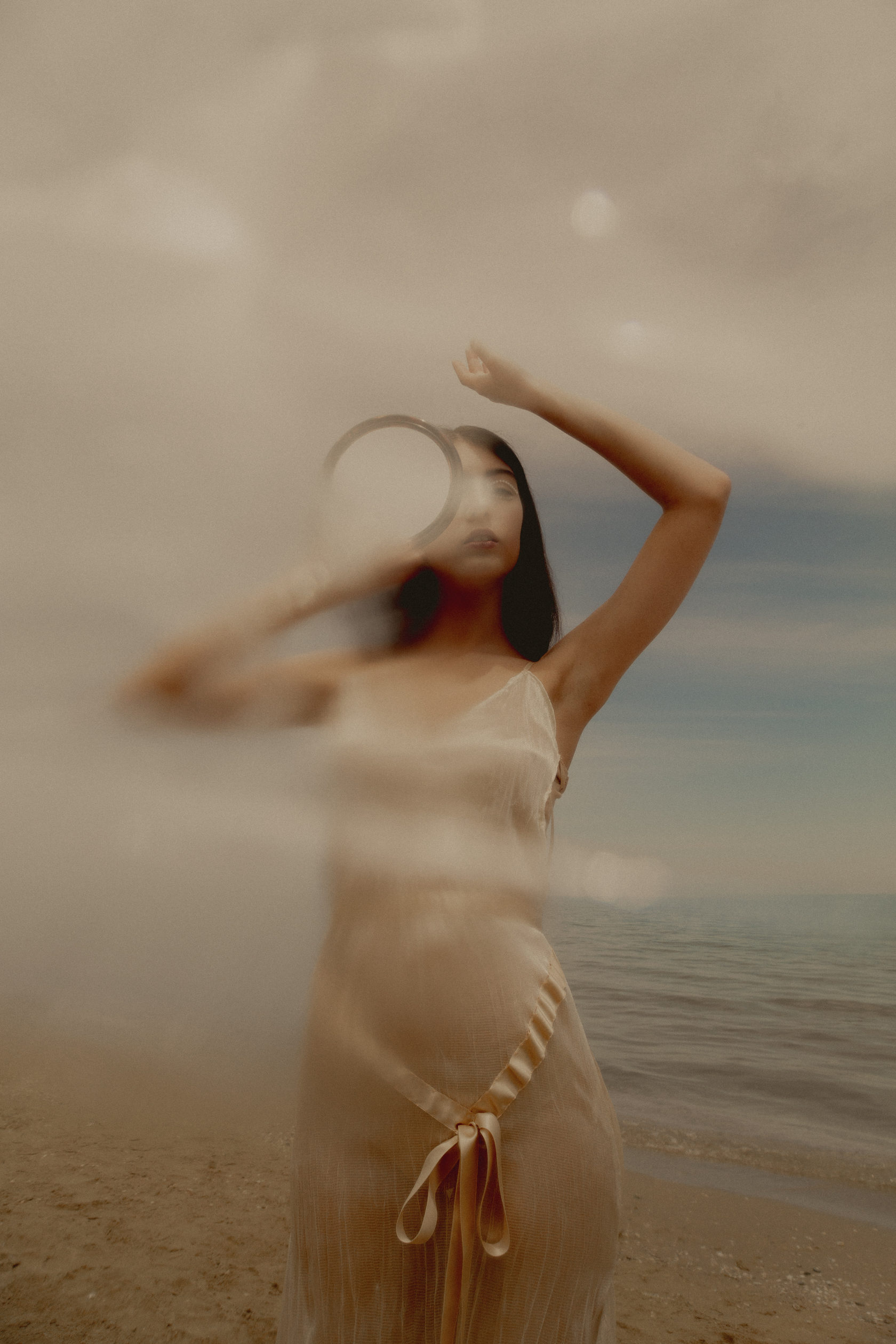Blurred figure of a woman in a flowing pale dress raising an arm near the ocean, bathed in soft warm golden light with a dreamy, atmospheric mood